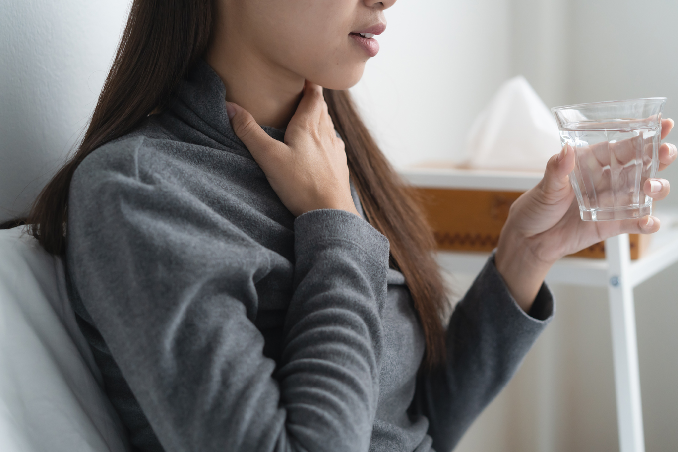 Close up of woman holding a glass of water and gripping her throat in pain on the sofa.