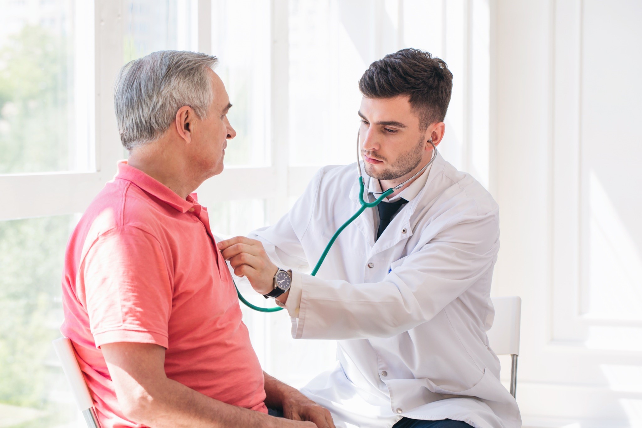 A male doctor is listening to the heart of an older male patient in an exam room.