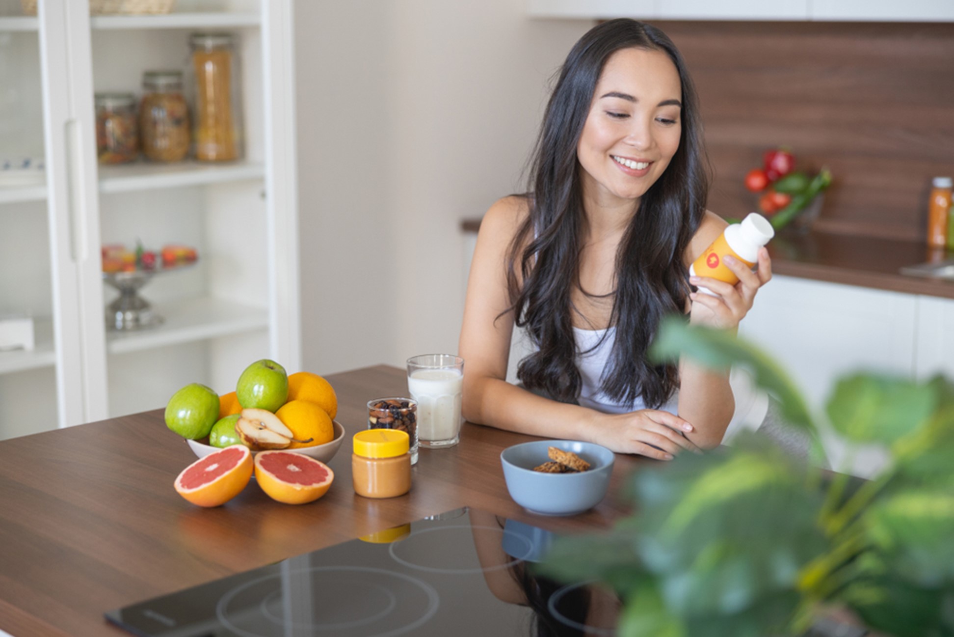 A woman smiling at a bottle of vitamins while sitting at her kitchen table next to fresh fruit.