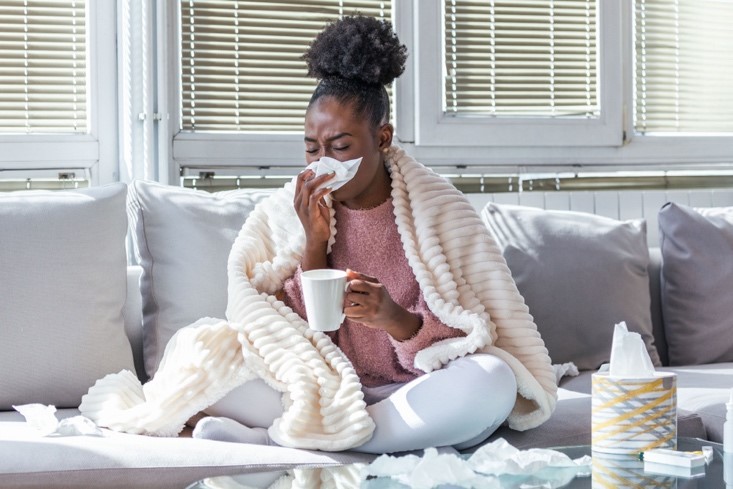 A sick African-American woman sitting on the sofa wrapped in a blanket, blowing her nose with one hand and holding a mug of tea in the other.