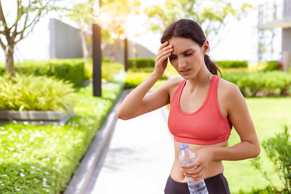 A woman in workout gear holds a water bottle in one hand with her other hand against her forehead in pain.