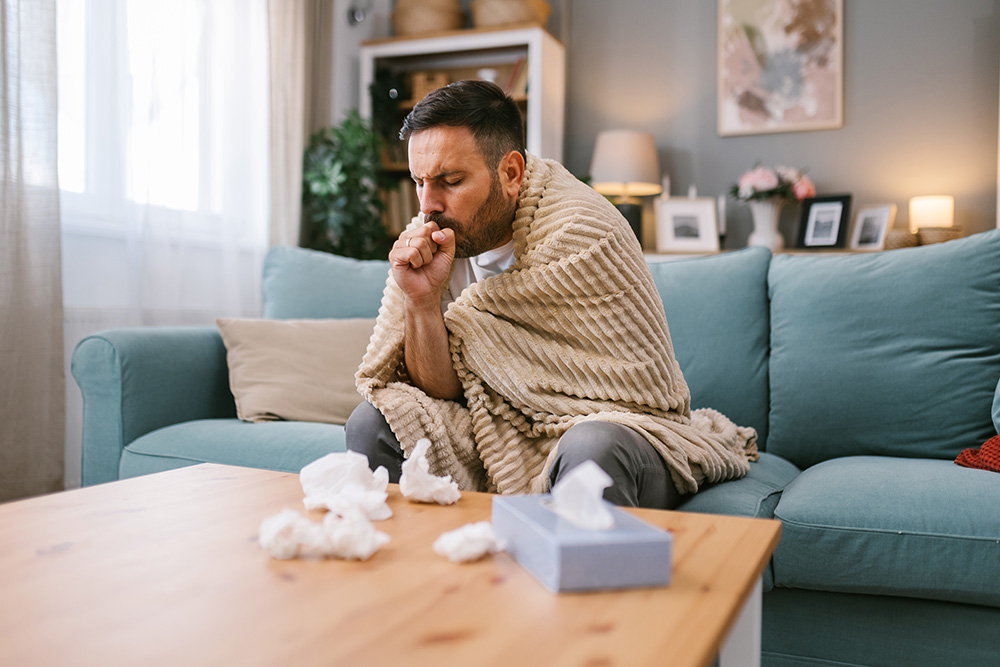A man sits on a couch wrapped in a blanket and appears to cough into his hand. Crumpled tissues are on the table in the foreground.