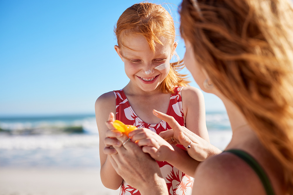 Little smiling red-haired girl and her mother applying sunscreen at the beach